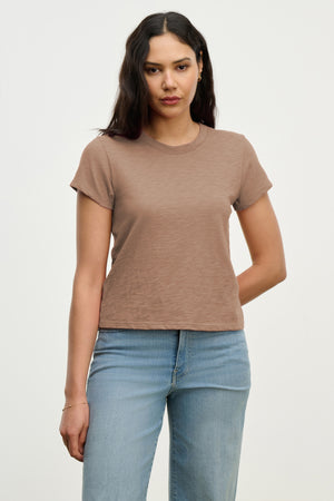 A woman with long dark hair wears the Velvet by Graham & Spencer MARIE TEE in brown and light blue jeans, standing against an off-white background.
