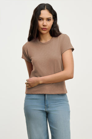 A woman with long dark hair wears the MARIE TEE by Velvet by Graham & Spencer in brown cotton slub, paired with light blue jeans, standing against a plain light background.