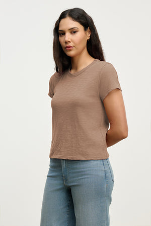 A woman wearing the MARIE TEE by Velvet by Graham & Spencer in light brown cotton slub and blue jeans stands against a plain light background with her hands behind her back.
