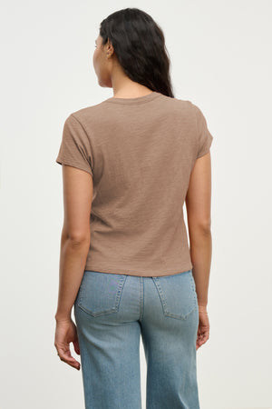 A woman stands facing away, wearing the MARIE TEE by Velvet by Graham & Spencer in brown with light blue jeans against a plain light background.