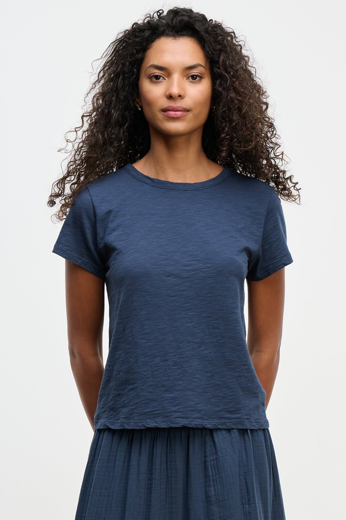   A person with curly hair, dressed in a relaxed boxy fit MARIE TEE from Velvet by Graham & Spencer and a matching navy skirt, poses against a plain background. Made from charming cotton slub material, this outfit is ideal for casual occasions. 