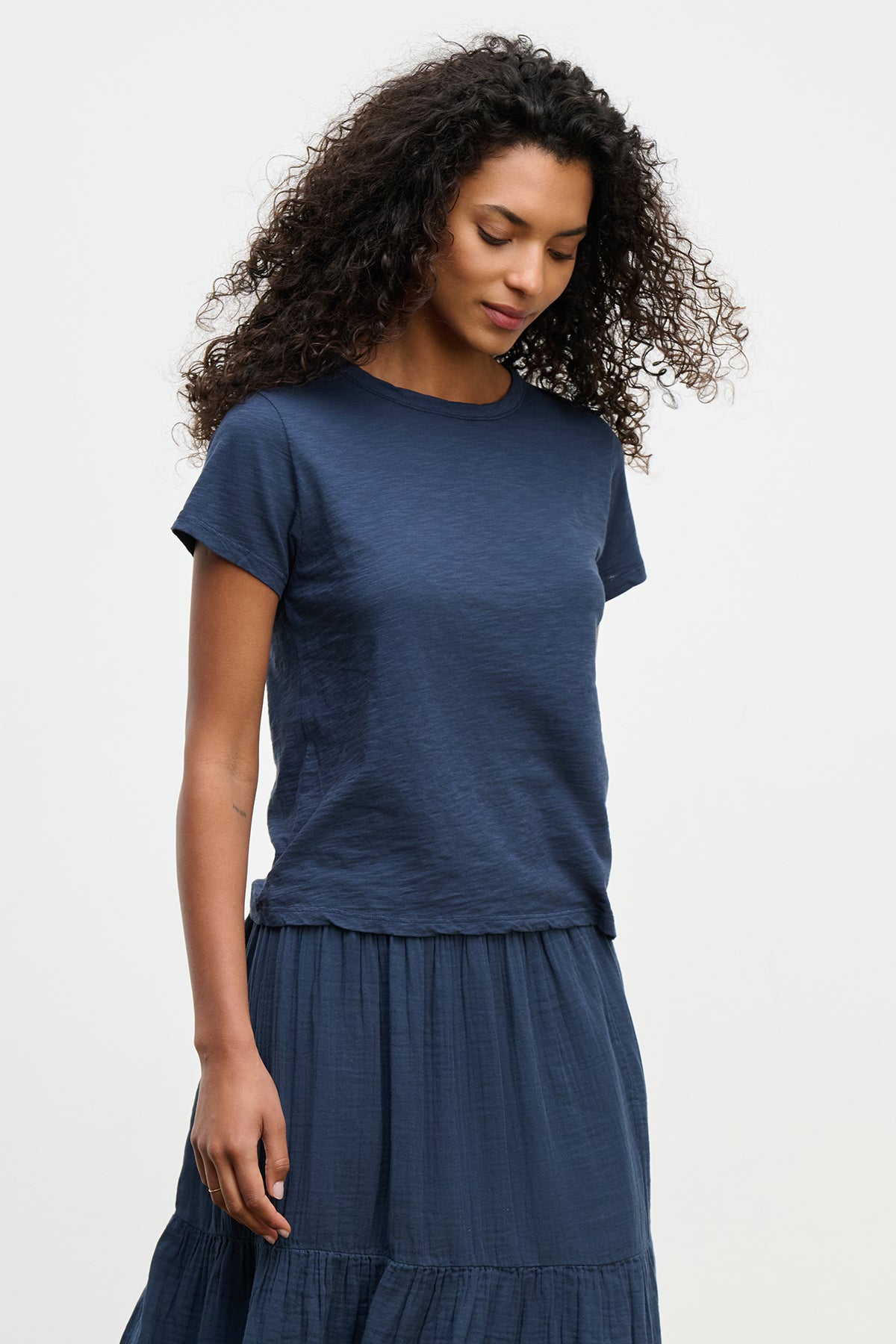   A person with curly hair stands against a plain background, wearing Velvet by Graham & Spencer's dark blue MARIE TEE and matching skirt. The cotton slub fabric ensemble offers a relaxed boxy fit, ideal for any casual occasion. 