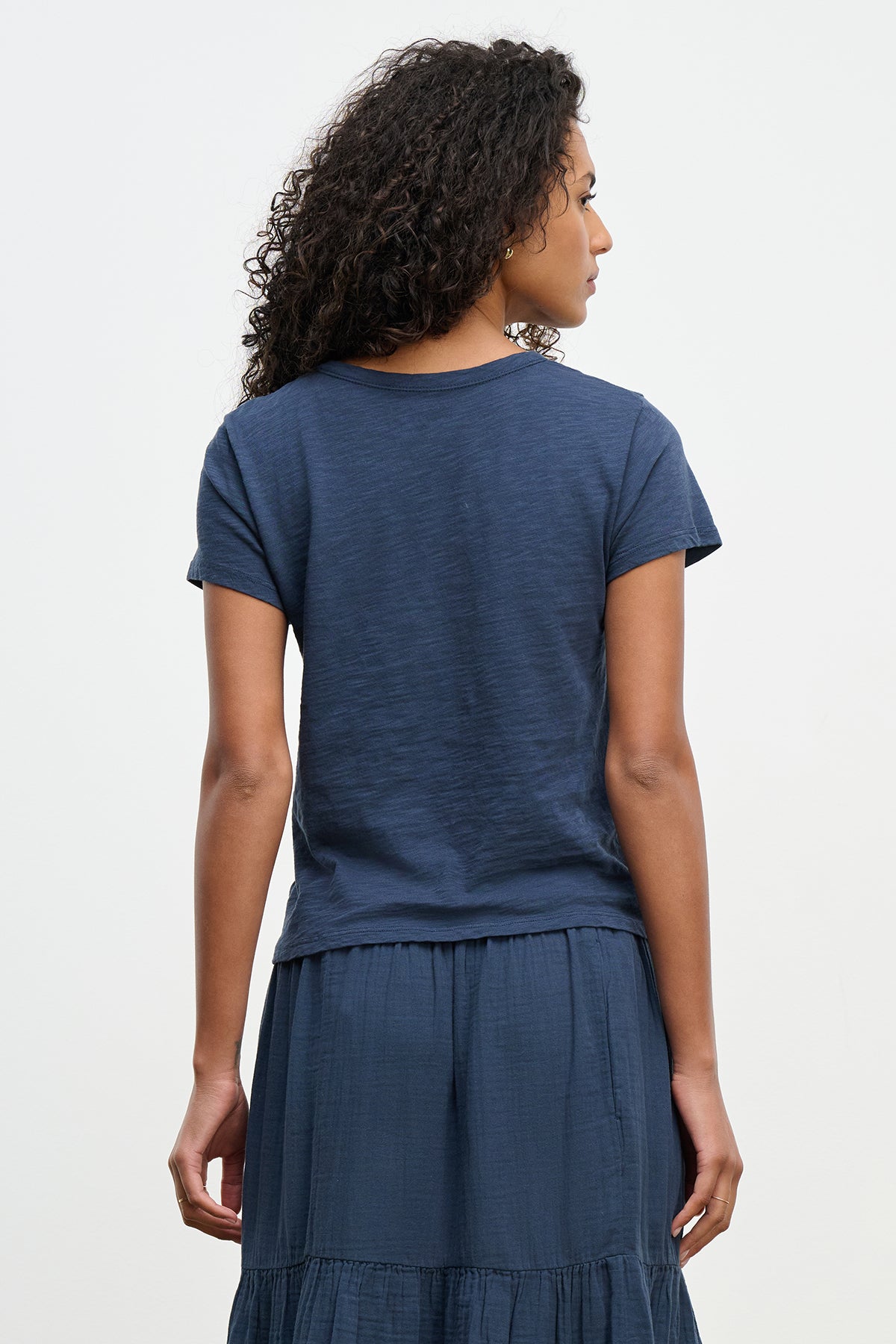   A person with curly hair is wearing the MARIE TEE by Velvet by Graham & Spencer, a navy blue relaxed boxy fit T-shirt and skirt, ideal for a casual occasion. 