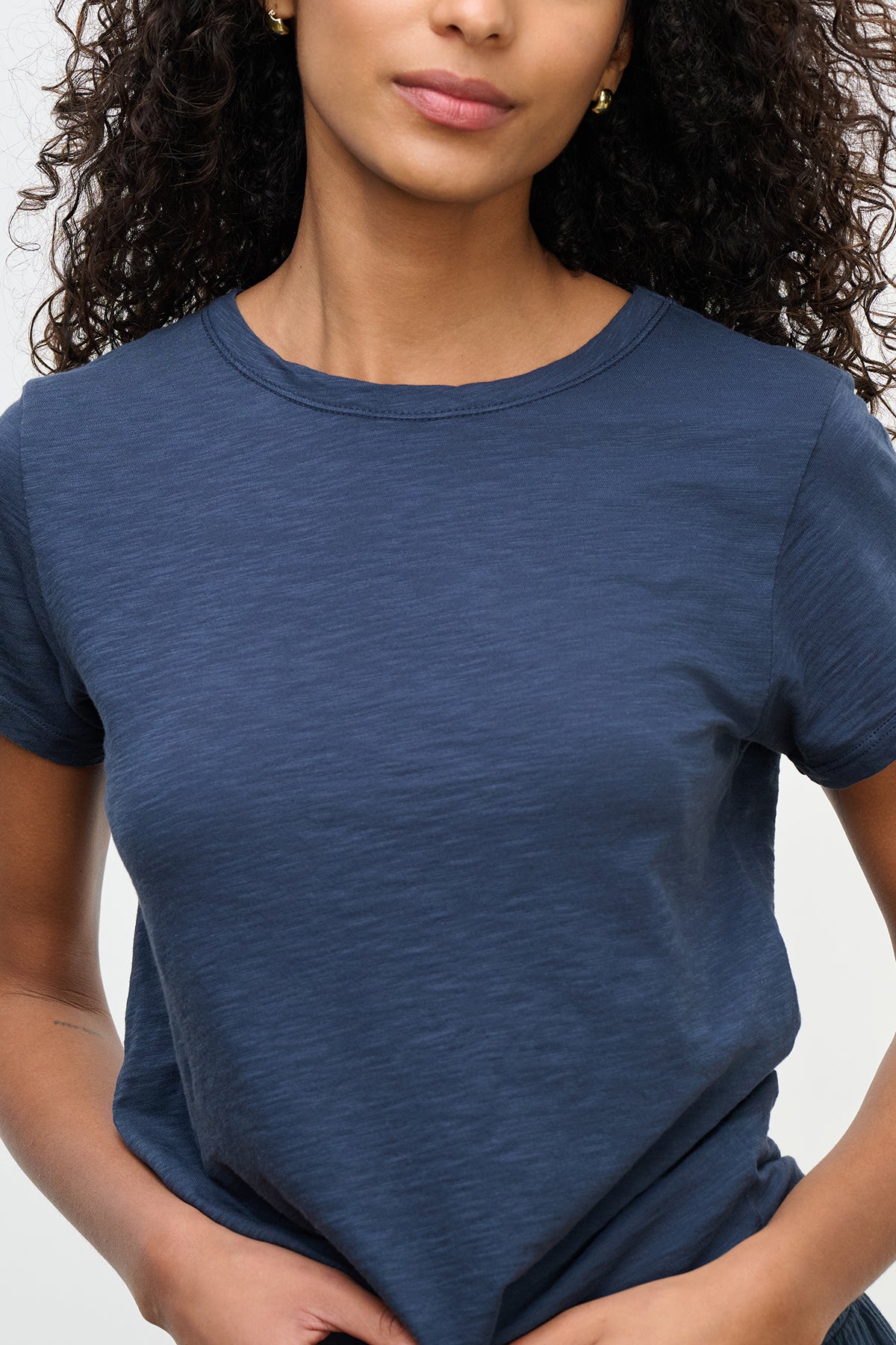   A person with curly hair wears the MARIE TEE by Velvet by Graham & Spencer, a cotton slub navy blue T-shirt featuring a relaxed boxy fit, against a plain background, ideal for casual occasions. 