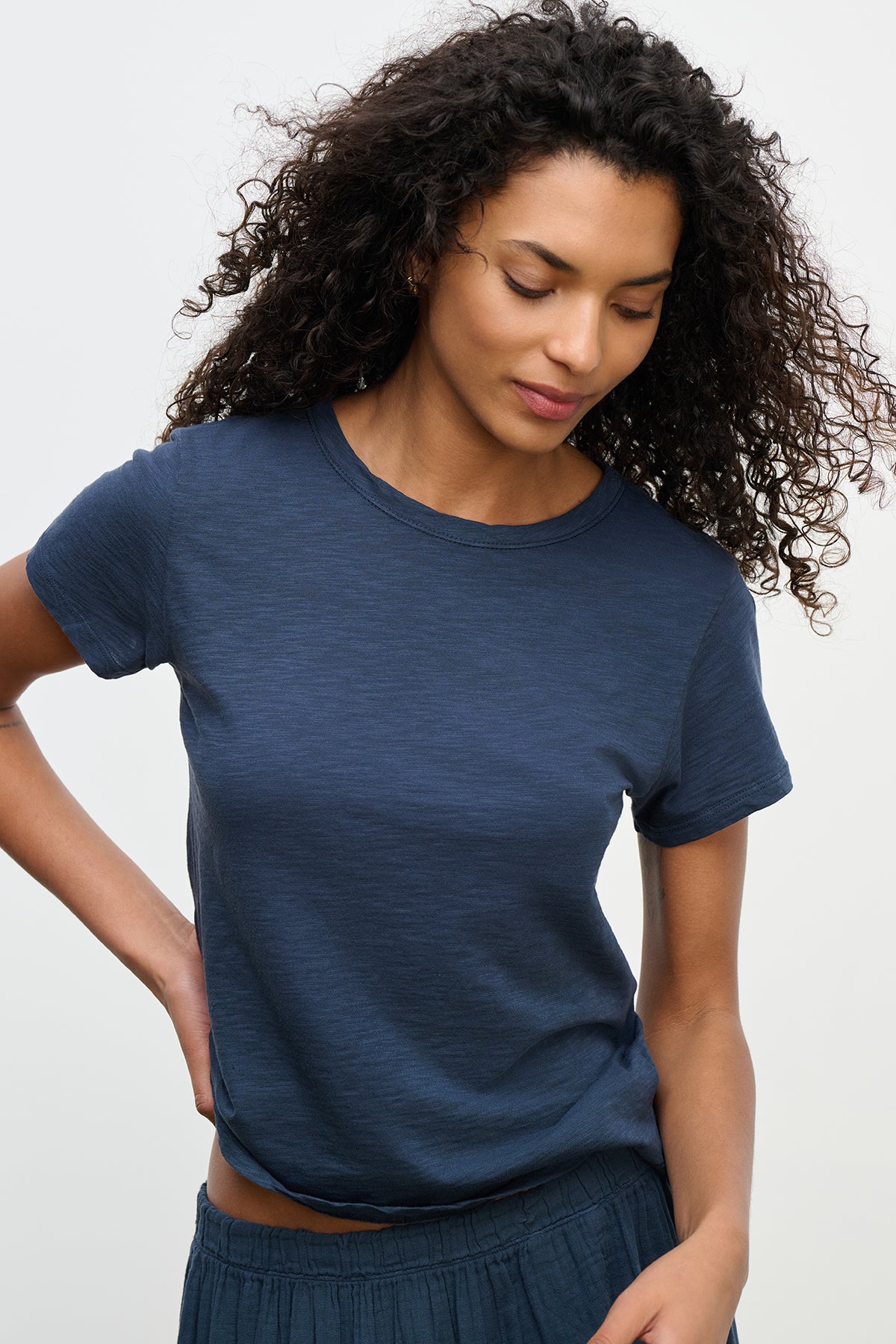 With curly hair, a person looks down with a neutral expression while wearing the MARIE TEE—a navy blue, relaxed boxy fit cotton slub t-shirt by Velvet by Graham & Spencer. This tee combines comfort and simplicity, making it perfect for casual occasions.-38662834520257