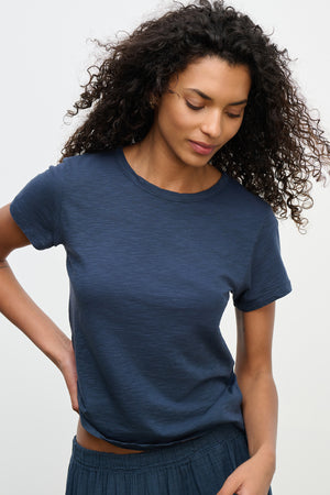 With curly hair, a person looks down with a neutral expression while wearing the MARIE TEE—a navy blue, relaxed boxy fit cotton slub t-shirt by Velvet by Graham & Spencer. This tee combines comfort and simplicity, making it perfect for casual occasions.