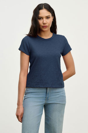 A woman with long dark hair wears the Velvet by Graham & Spencer MARIKA TEE in navy, a relaxed boxy cotton slub style, paired with light blue jeans and standing against a plain light background.
