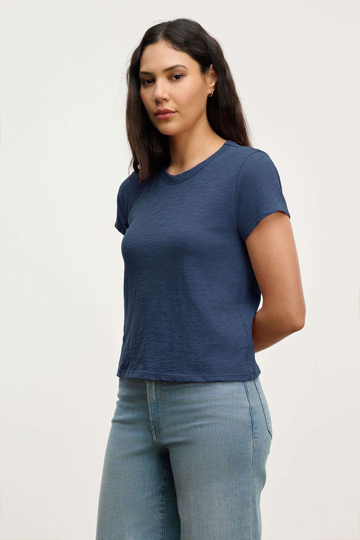   A woman with long dark hair wears the Velvet by Graham & Spencer MARIKA TEE, a navy cotton slub top in a relaxed boxy fit, paired with light blue jeans and standing against a plain light background with her arms behind her back. 