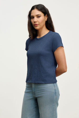 A woman with long dark hair wears the Velvet by Graham & Spencer MARIKA TEE, a navy cotton slub top in a relaxed boxy fit, paired with light blue jeans and standing against a plain light background with her arms behind her back.