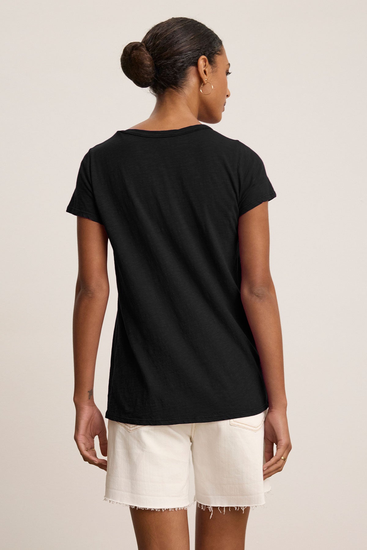 A person stands with their back to the camera, wearing the TILLY TEE by Velvet by Graham & Spencer, a classic crew neck made from soft cotton slub, paired with white shorts against a plain background.-39145410560193