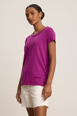 A woman in a purple TILLY TEE by Velvet by Graham & Spencer and white frayed hem shorts stands against a plain light background.