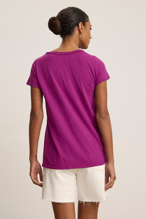 A person stands facing away, wearing the TILLY TEE by Velvet by Graham & Spencer in purple with white shorts. Their hair is tied back in a neat bun.