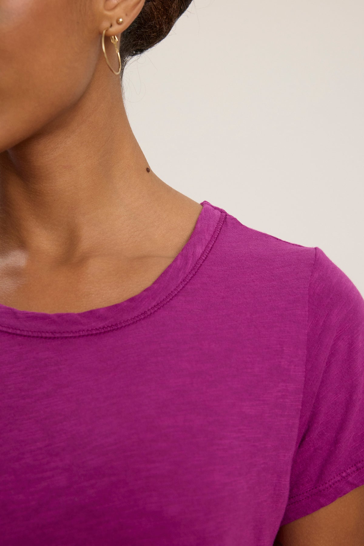 Close-up of a person wearing the TILLY TEE by Velvet by Graham & Spencer in purple cotton slub, styled with a gold hoop earring. Part of their face and shoulder is visible.-39111856128193