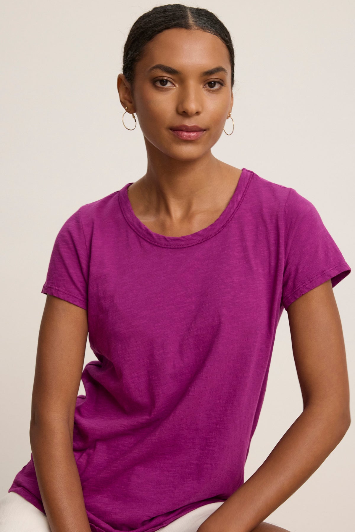 A woman with short, dark hair sits against a plain background, wearing the bright purple TILLY TEE by Velvet by Graham & Spencer and gold hoop earrings.-39111856193729