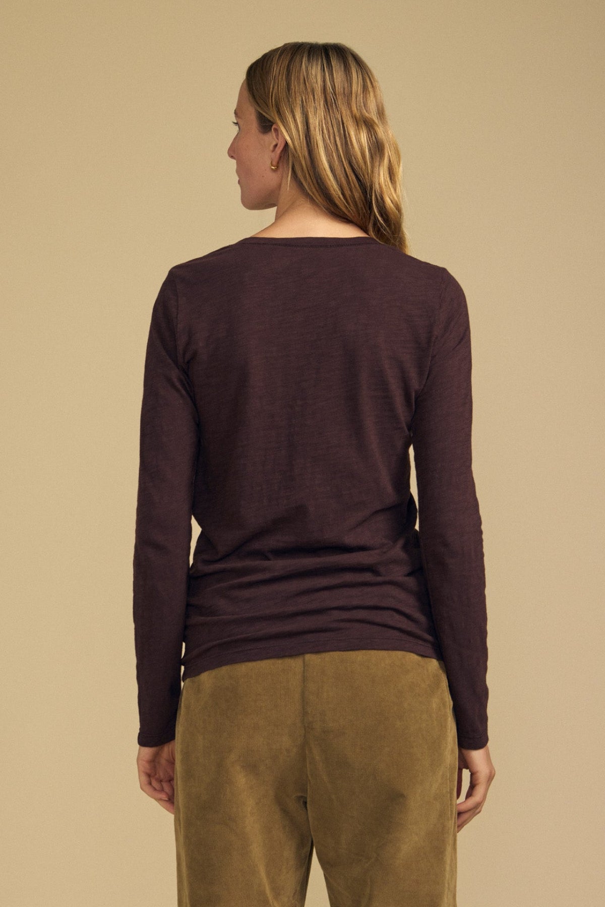 A person stands facing away, wearing the Velvet by Graham & Spencer BLAIRE TEE—a long-sleeve dark brown cotton slub top with a subtle v-neckline—paired with olive green pants against a plain beige background.
