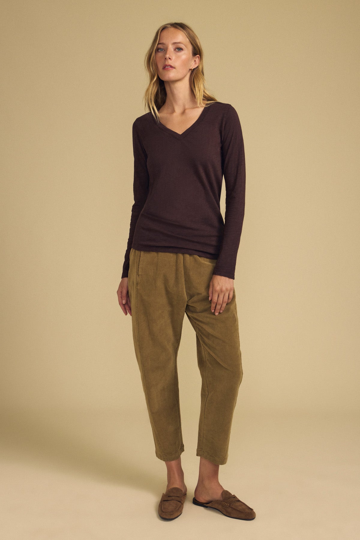 A person stands against a beige background wearing the BLAIRE TEE by Velvet by Graham & Spencer—a long sleeve, v-neckline cotton slub tee in brown—paired with olive pants and brown slip-on shoes.