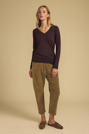 A person stands against a beige background wearing the BLAIRE TEE by Velvet by Graham & Spencer—a long sleeve, v-neckline cotton slub tee in brown—paired with olive pants and brown slip-on shoes.