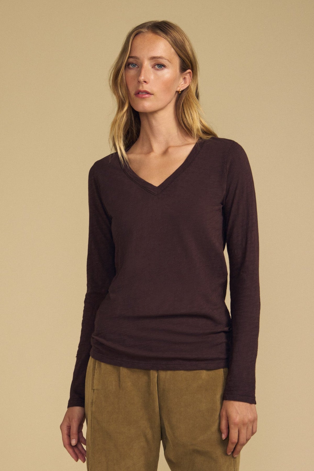 A person stands against a beige background, wearing the BLAIRE TEE by Velvet by Graham & Spencer—a long sleeve dark brown V-neckline cotton slub top—paired with light brown pants.