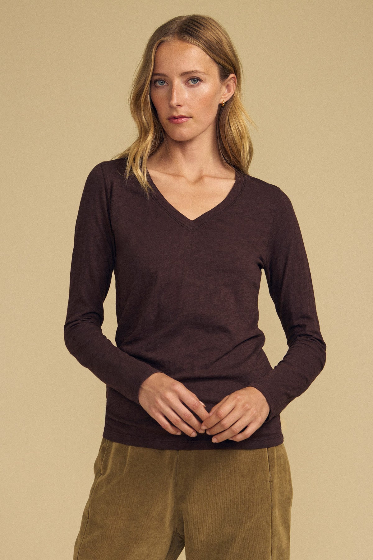 A woman stands against a beige background wearing the BLAIRE TEE by Velvet by Graham & Spencer—a dark brown, long sleeve, V-neckline cotton slub tee—paired with light brown pants, her hands loosely clasped in front of her.-42063726936257