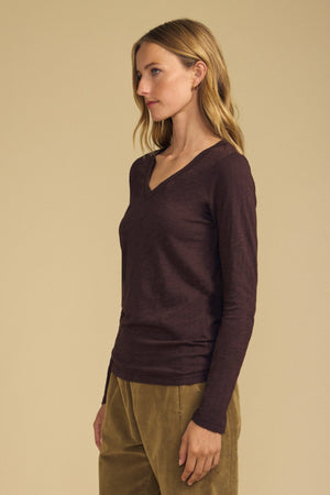 A woman stands in profile against a beige background, wearing the Velvet by Graham & Spencer BLAIRE TEE—a long sleeve, dark brown V-neckline cotton slub top—paired with tan pants.