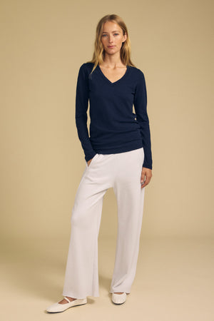 A woman stands against a beige background wearing the BLAIRE TEE by Velvet by Graham & Spencer—a navy long sleeve V-neckline cotton slub top—paired with white wide-leg pants and white ballet flats.