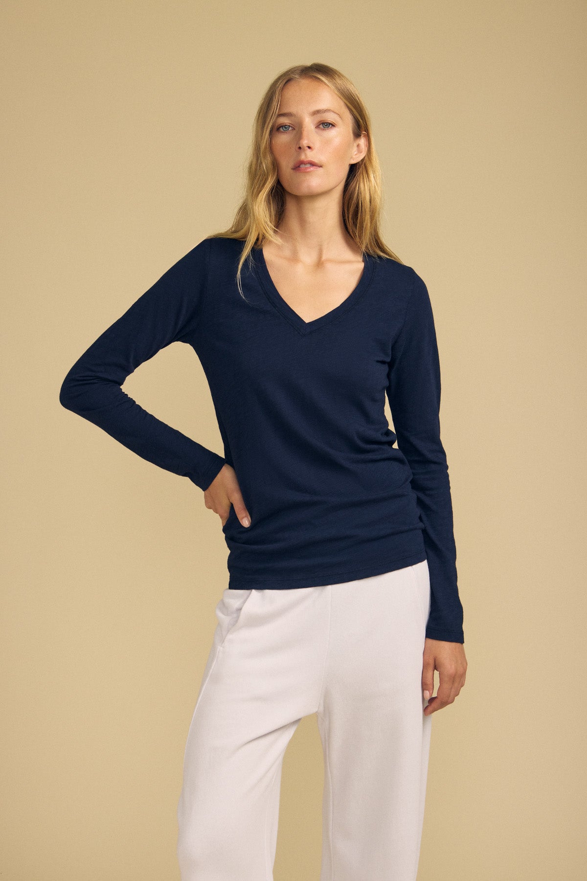 A person wearing the BLAIRE TEE by Velvet by Graham & Spencer—a navy blue, long sleeve cotton slub V-neck top—paired with white pants stands against a plain beige background.