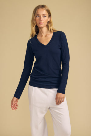 A woman stands against a beige background wearing the BLAIRE TEE by Velvet by Graham & Spencer, a navy cotton slub V-neck with long sleeves, paired with white pants and displaying a neutral expression.