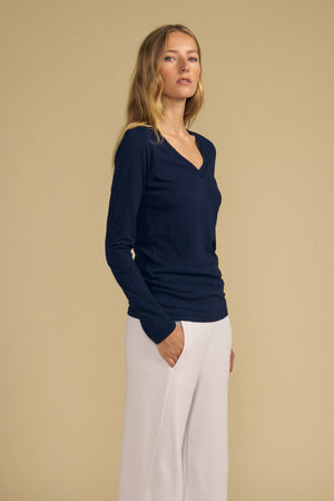 A woman stands against a beige background, wearing the BLAIRE TEE by Velvet by Graham & Spencer—a navy cotton slub long sleeve top featuring a flattering v-neckline—paired with white pants and one hand in her pocket.