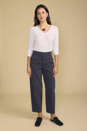 A woman stands against a beige background wearing a white long-sleeve shirt, black shoes, a red pendant necklace, and the TENLEY PANT in gray by Velvet by Graham & Spencer.