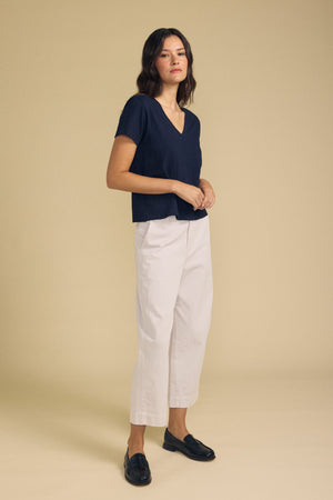A person stands against a plain beige background, wearing the ELISHA TEE by Velvet by Graham & Spencer—a navy V-neck made from soft cotton slub—paired with white cropped pants and black loafers for a timeless wardrobe staple.
