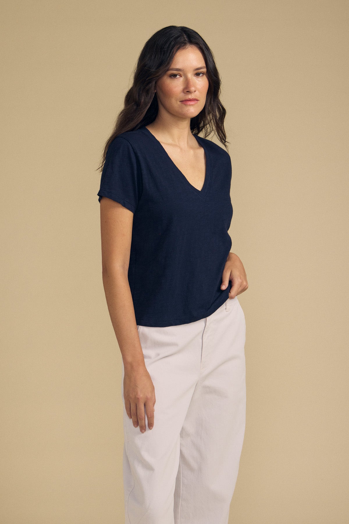 A woman with long brown hair stands against a beige background wearing the ELISHA TEE by Velvet by Graham & Spencer—a navy cotton slub V-neck paired with light-colored pants for a versatile, timeless look.-41798258524353