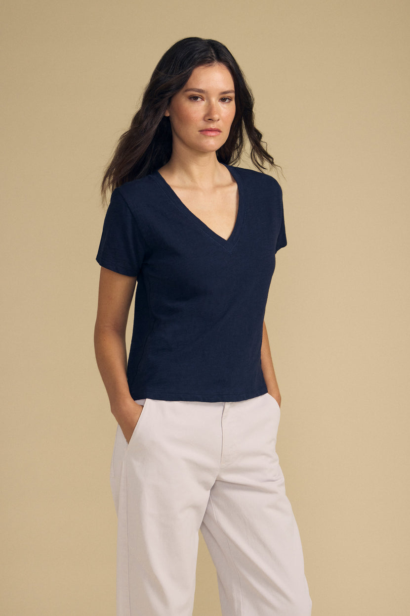 Woman standing against a beige background, wearing the ELISHA TEE by Velvet by Graham & Spencer—a short-sleeved navy cotton slub V-neck tee—paired with light-colored pants and hands in pockets: a timeless wardrobe essential.