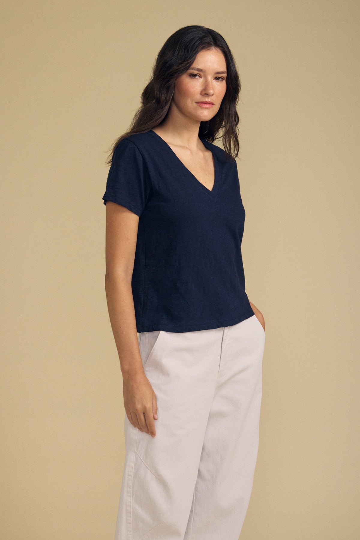 A woman with long dark hair stands against a plain beige background, one hand in her pocket, wearing the ELISHA TEE by Velvet by Graham & Spencer—a navy cotton slub V-neck tee—paired with light-colored pants.-41798258458817