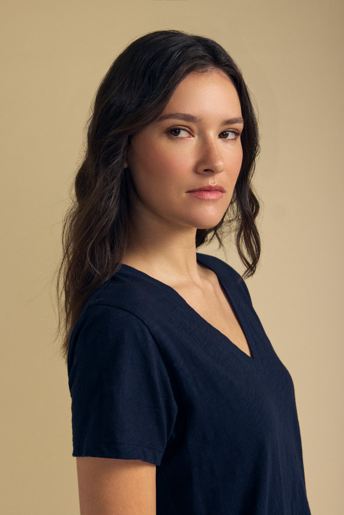 A woman with long brown hair wearing the ELISHA TEE by Velvet by Graham & Spencer—a classic V-neck crafted from soft cotton slub—stands against a plain beige background, gazing slightly to the side with a neutral expression.
