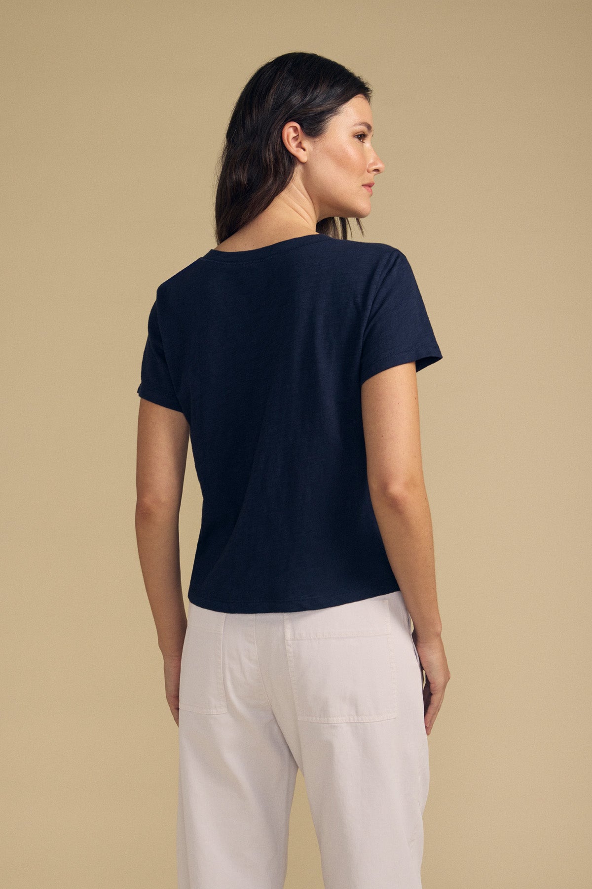 A woman stands facing away, wearing the Velvet by Graham & Spencer ELISHA TEE—a short-sleeved navy blue V-neck—paired with white pants against a plain beige background, showcasing this timeless wardrobe staple.