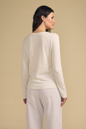 A woman with long dark hair is seen from behind, standing against a beige background, wearing the Velvet by Graham & Spencer LIZZIE TEE—a long sleeve crew neck tee—paired with light pants.