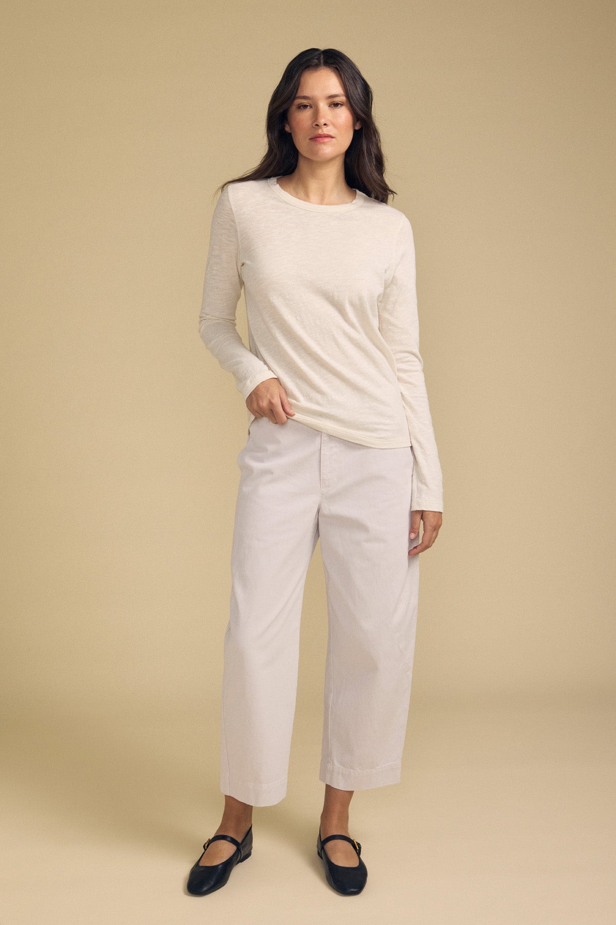 A woman stands against a beige background, wearing the LIZZIE TEE by Velvet by Graham & Spencer—a long-sleeve cream textured cotton slub top with a classic crew neckline—paired with wide-leg white pants and black Mary Jane shoes.-41925583569089