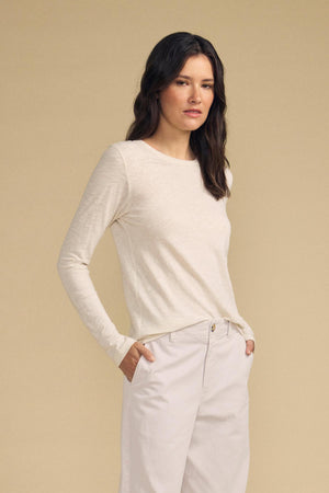 A woman with long dark hair wears the LIZZIE TEE by Velvet by Graham & Spencer—a cream-colored long sleeve tee with a classic crew neckline—paired with light beige pants, standing against a plain beige background.
