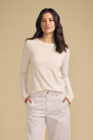 A woman with long dark hair stands against a plain beige background with her hands in her pockets, wearing the LIZZIE TEE by Velvet by Graham & Spencer—a light beige long sleeve tee featuring a classic crew neckline—paired with light pants.