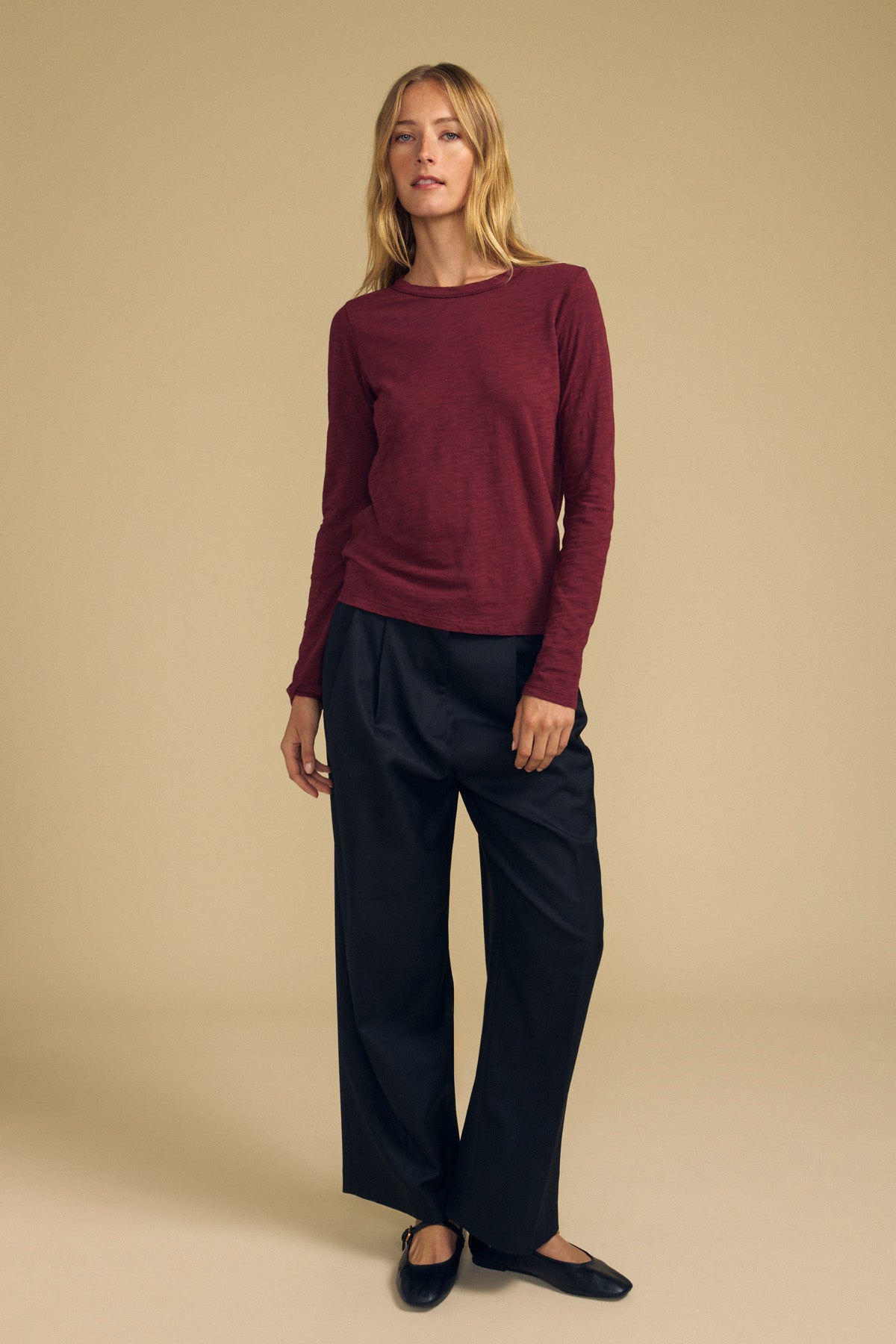 A woman stands against a beige background, wearing the LIZZIE TEE by Velvet by Graham & Spencer—a long-sleeve maroon crew neck top—paired with black wide-leg pants and black flats.