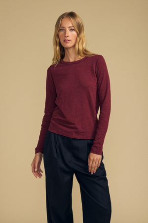 A person stands against a beige background, wearing the LIZZIE TEE—a long sleeve cotton slub crew neck tee in maroon—by Velvet by Graham & Spencer, paired with dark loose-fitting pants.