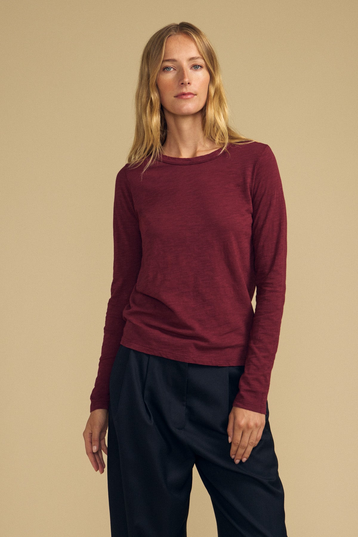 A person with long blond hair wears the Velvet by Graham & Spencer LIZZIE TEE, a maroon textured cotton slub long-sleeve shirt with a classic crew neckline, paired with black pants, standing against a plain beige background.
