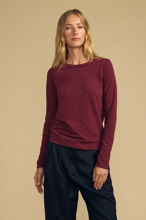 A person with long blond hair wears the Velvet by Graham & Spencer LIZZIE TEE, a maroon textured cotton slub long-sleeve shirt with a classic crew neckline, paired with black pants, standing against a plain beige background.