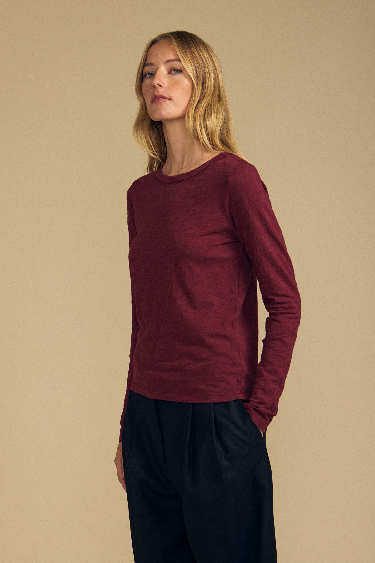 A person with long blonde hair wears the Velvet by Graham & Spencer LIZZIE TEE, a maroon cotton slub top featuring long sleeves and a classic crew neckline, paired with black pants and standing against a plain beige background.