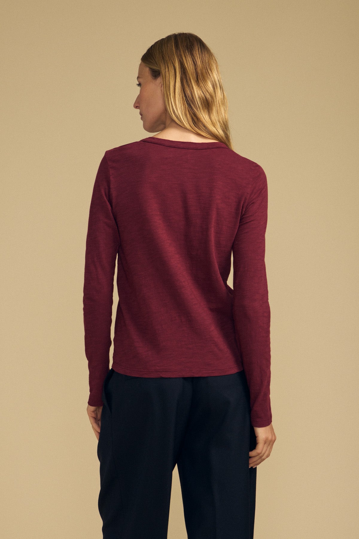 A person stands with their back to the camera, wearing the LIZZIE TEE by Velvet by Graham & Spencer—a long-sleeve maroon crew neck cotton slub top—paired with dark pants, set against a plain beige background.-41964606554305
