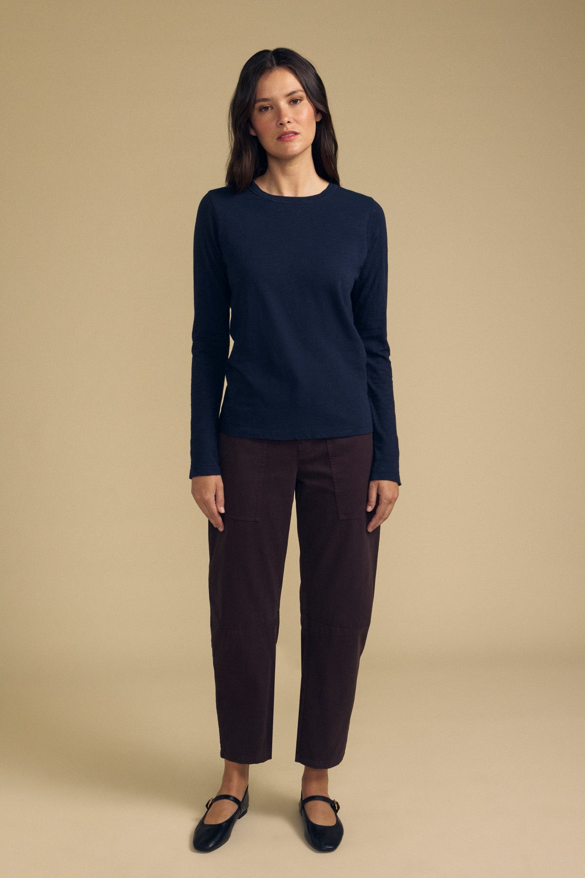 A woman stands against a beige background wearing the LIZZIE TEE by Velvet by Graham & Spencer, a long sleeve crew neck top in navy textured cotton slub, paired with dark brown cropped pants and black flat shoes.