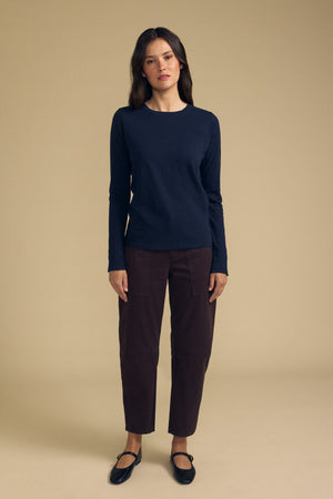 A woman stands against a beige background wearing the LIZZIE TEE by Velvet by Graham & Spencer, a long sleeve crew neck top in navy textured cotton slub, paired with dark brown cropped pants and black flat shoes.