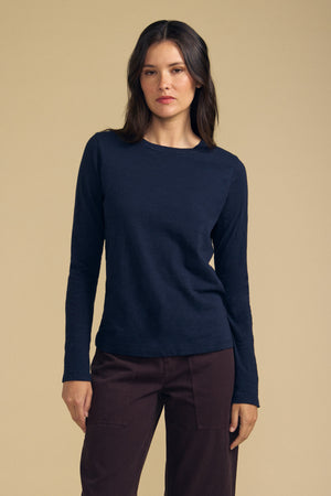 A woman with dark hair is wearing the Velvet by Graham & Spencer LIZZIE TEE in navy, featuring long sleeves and a classic crew neckline, paired with brown pants and standing against a beige background.