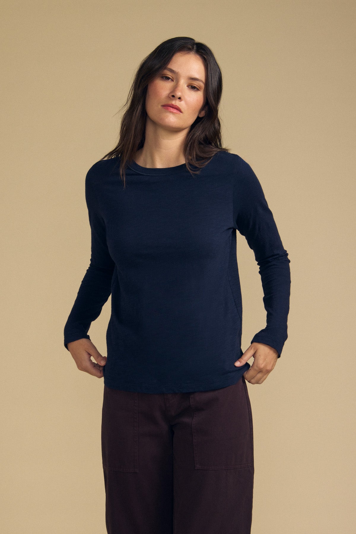 A person with long brown hair wears the LIZZIE TEE by Velvet by Graham & Spencer, a navy long sleeve top with a classic crew neckline, paired with dark pants and standing against a plain beige background.
