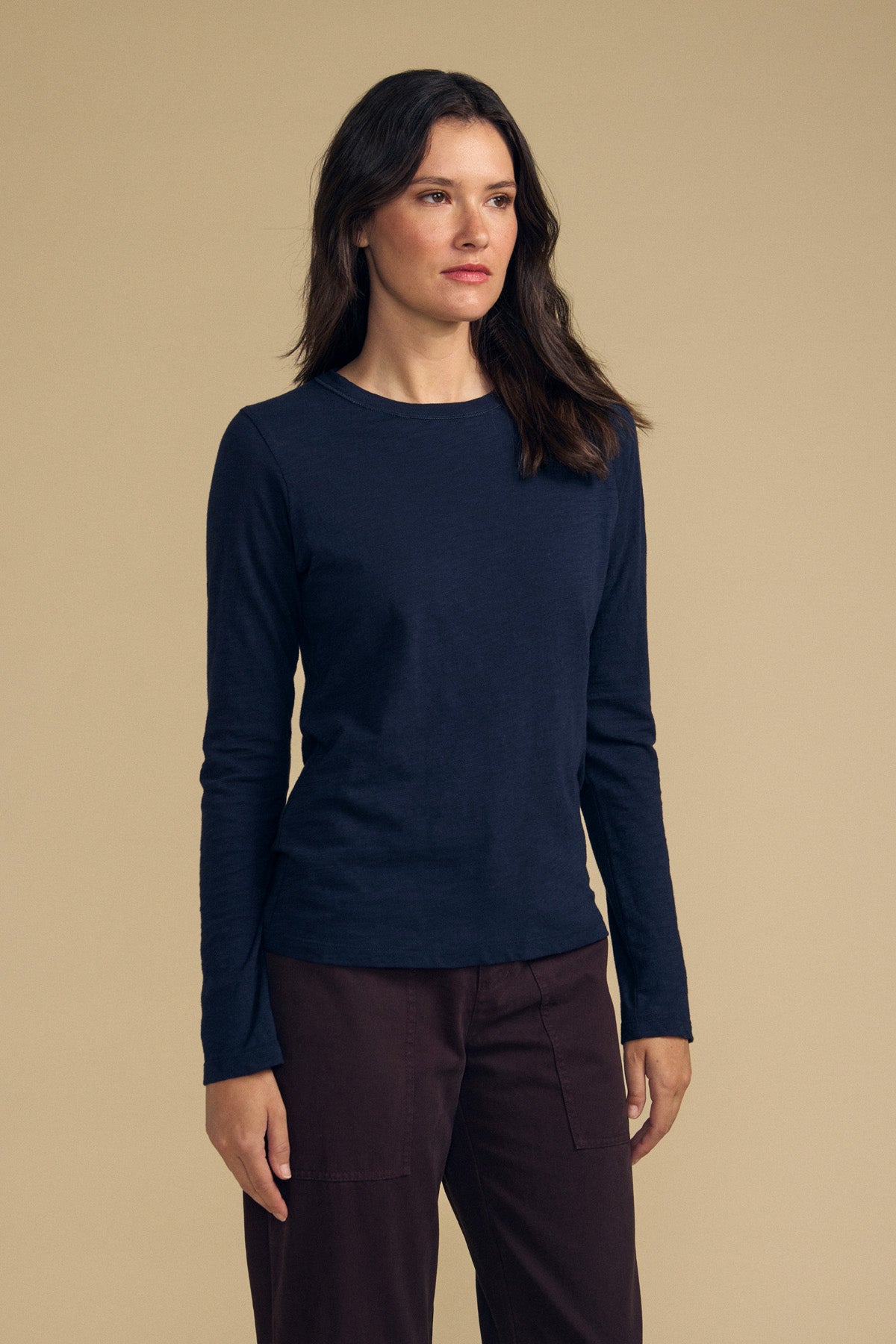 A woman stands against a beige background, wearing the LIZZIE TEE by Velvet by Graham & Spencer—a navy long-sleeve top with a classic crew neckline—paired with dark brown pants, her arms relaxed at her sides.